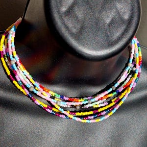 Op de afbeelding: Een meerstrengs kralenketting met verschillende kleuren, waaronder roze, geel, rood, blauw en groen. De ketting is gemaakt van kleine, ronde kralen en is gelaagd op een zwart manikin.