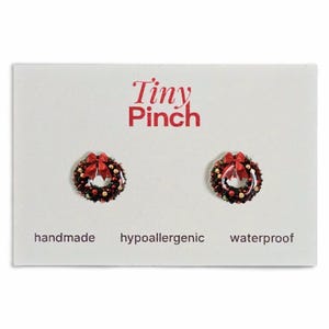 May include: A pair of Christmas wreath stud earrings with red bows, displayed on a white card. The card has the text "Tiny Pinch", "handmade", "hypoallergenic", and "waterproof". The earrings are festive and suitable for holiday wear.