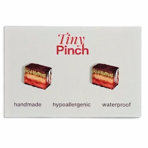 May include: A pair of miniature cake earrings on a white card with the text "Tiny Pinch" in red. The earrings are shaped like layered cakes with red, tan, and brown colors. The card also says "handmade", "hypoallergenic", and "waterproof".