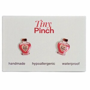 Könnte beinhalten: Ein Paar herzförmige Ohrringe mit rotem Trank-Design, beschriftet mit "Love Potion" auf einer weißen Karte. Die Karte enthält auch den Text "Tiny Pinch", "handmade", "hypoallergenic" und "waterproof".