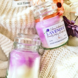 May include: Two glass jars of lavender scented candles with a white and purple ombre effect. The label on the jar reads "Calming & reduces anxiety LAVENDER Promotes feeling of happiness" and "HAND-POURED IN DENMARK".