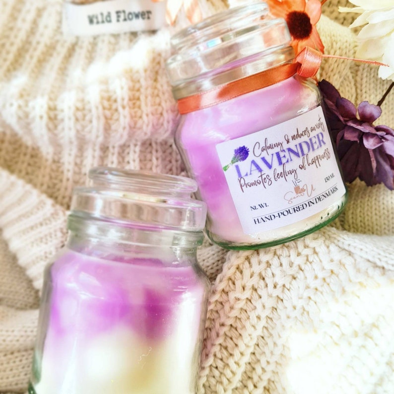 May include: Two glass jars of lavender scented candles with a white and purple ombre effect. The label on the jar reads "Calming & reduces anxiety LAVENDER Promotes feeling of happiness" and "HAND-POURED IN DENMARK".