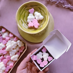 Puede incluir: Unas ceras para fundir en forma de corazón, de color blanco y rosa, en una pequeña caja blanca abierta. Las ceras para fundir son visibles y de formas variadas, incluyendo corazones y nubes.