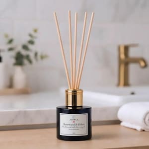 Op de afbeelding: Een zwarte glazen rietdiffuserfles met een goudkleurige kraag en vijf lichtgekleurde houten rietjes. Het witte etiket bevat de tekst SCENT.U, Rosewood & Velvet en 100ml. De fles heeft een cilindrische basis en een smalle hals voor de rietjes.