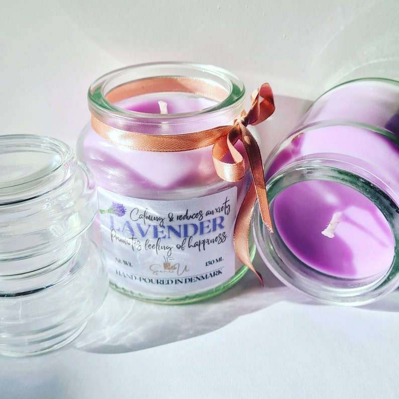 May include: Two glass jars with lavender scented candles. The jars are clear glass with a purple candle inside. The candle in the front jar has a gold ribbon tied around it. The label on the front jar reads "Calming & reduces anxiety Lavender promotes feeling of happiness. 150 ml. Hand-poured in Denmark."