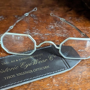 May include: Silver-toned metal spectacles with octagonal lenses and thin, hinged wire temples. The glasses rest on a black leather case featuring the gold-printed text: VAIL, AZ & LIVINGSTON, NJ, Historic EyeWear Co., THOS. VALENZA OPTICIAN.