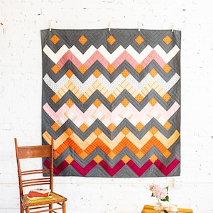 May include: A gray chevron quilt with pink, orange, and yellow geometric patterns hanging on a white brick wall. A wooden chair with a stack of books sits in front of the quilt. A wooden crate with a vase of pink flowers sits on the floor.