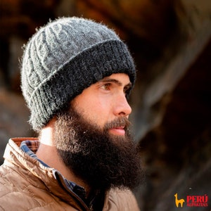 Op de afbeelding: Een man met een baard draagt een grijze en zwarte gebreide beanie. De beanie heeft een kabelbreipatroon en een omslagrand. De man kijkt naar rechts.