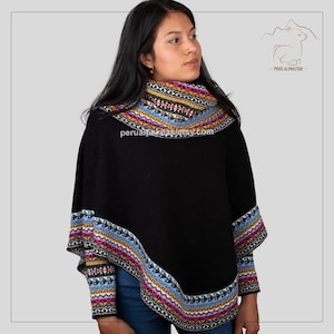 May include: Black alpaca wool poncho with colourful geometric patterns around the neckline and hem. The poncho has long sleeves with colourful geometric patterns on the cuffs. The model is wearing blue jeans.