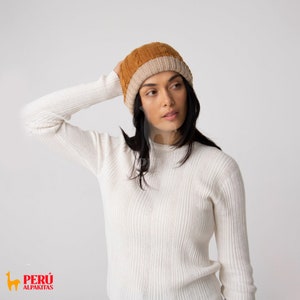 Op de afbeelding: Een vrouw draagt een witte geribbelde trui en een bruin en beige gebreide muts.