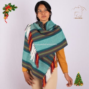 Puede incluir: Un chal de lana de alpaca de color verde azulado y blanco con flecos coloridos. El chal tiene un patrón geométrico y lo lleva puesto una persona con una camisa amarilla.