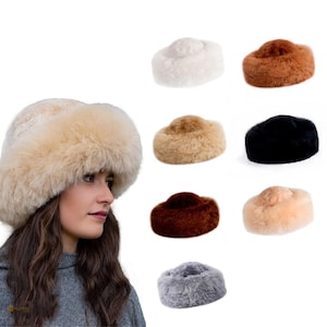 May include: A woman wearing a beige fur hat, with a selection of similar hats in various colours. The hats are round with a fluffy texture, and the additional hats are white, brown, black, and grey.