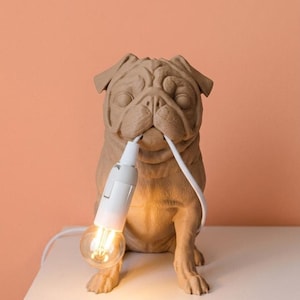 Puede incluir: Una lámpara de mesa con forma de pug de color marrón claro y acabado mate. La figura del perro sostiene en su boca un cable eléctrico blanco, conectado a un casquillo blanco y a una bombilla LED de cristal transparente que emite luz cálida.