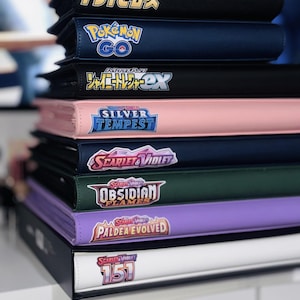 May include: A stack of nine black, blue, pink, green, purple, and white binders with various Pokemon card game logos, including "Pokemon Go", "Silver Tempest", "Scarlet & Violet", and "Obsidian Flames".
