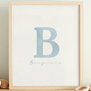 Puede incluir: Una impresión enmarcada con una letra "B" azul claro y el nombre "Benjamin" en escritura delicada. Un oso de peluche y un sonajero de madera están sobre una superficie de madera, sugiriendo un tema de guardería.
