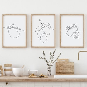 Könnte beinhalten: Drei minimalistische Line-Art-Drucke in Holzrahmen, die eine Zitrone, einen Apfel und eine Erdbeere zeigen. Die Drucke hängen an einer weißen Wand über einer Holzarbeitsplatte mit einem Waschbecken und einem Wasserhahn.
