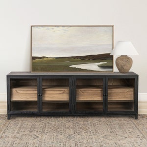 May include: A black cabinet with glass doors and wooden drawers, topped with a framed landscape painting and a lamp with a white shade. A patterned rug is on the floor.