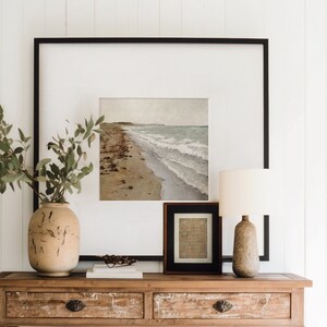 May include: A wooden console table with two drawers and a lower shelf. The table is decorated with a vase of greenery, a framed seascape print, a framed print, and a lamp.