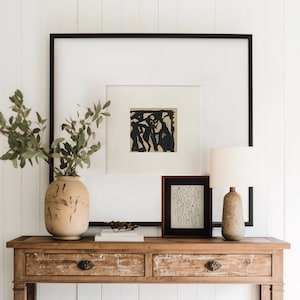 May include: A wooden console table with two drawers and a lower shelf. The table is decorated with a vase of greenery, a framed black and white abstract print, a framed black and white line drawing, and a ceramic lamp with a cream-colored shade.