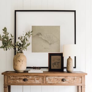 Puede incluir: Una consola de madera con dos cajones y un estante inferior. La mesa está decorada con un jarrón de vegetación, una impresión enmarcada de un paisaje de montaña y una ilustración botánica enmarcada. Una lámpara de mesa de color crema se encuentra sobre la mesa.