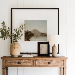 Peut inclure: Une console en bois avec deux tiroirs et une étagère inférieure. La table est décorée d'un vase de verdure, d'une peinture de paysage encadrée, d'un dessin encadré et d'une lampe avec un abat-jour blanc.