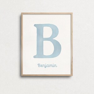 May include: A framed watercolour print displaying a large, pale blue letter "B" on a white backdrop. The name "Benjamin" is written in a matching blue script beneath the letter. The frame is made of light-coloured wood.
