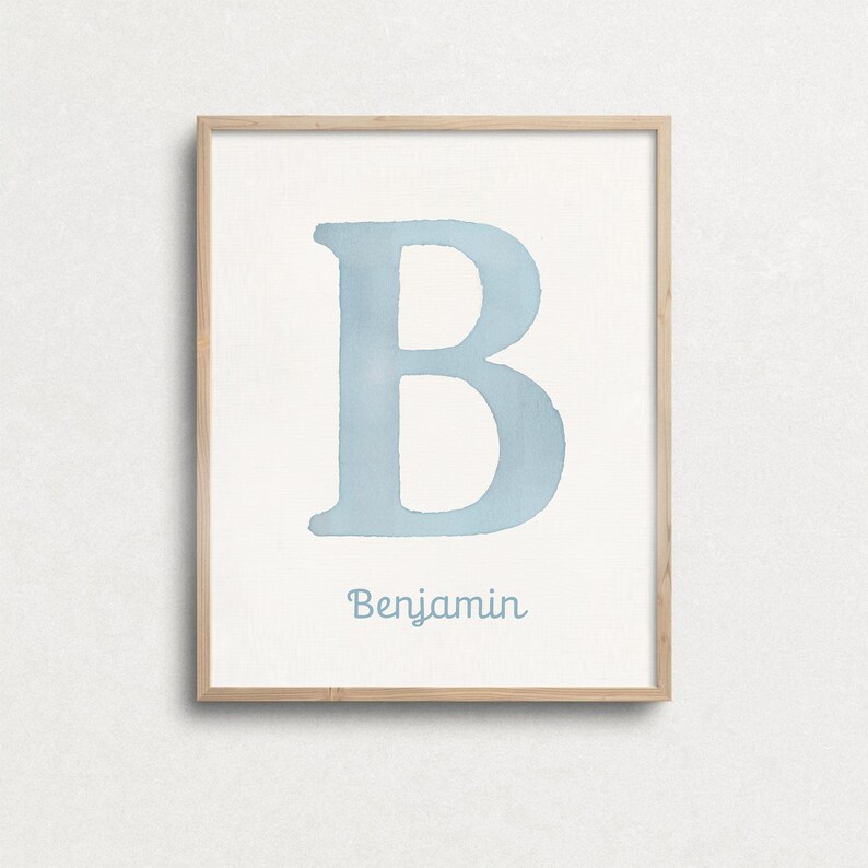 May include: A framed watercolour print displaying a large, pale blue letter "B" on a white backdrop. The name "Benjamin" is written in a matching blue script beneath the letter. The frame is made of light-coloured wood.