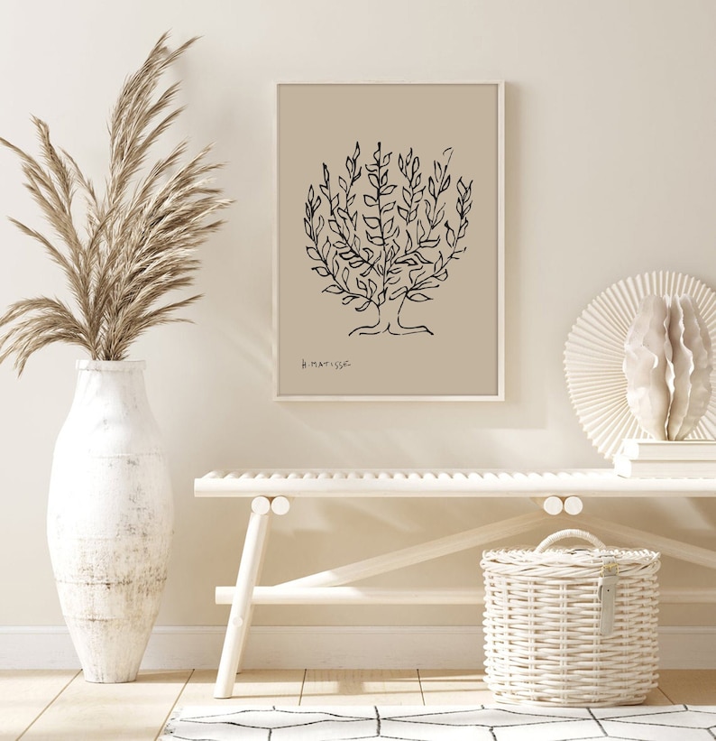 May include: A black and white line drawing of a tree on a beige background. The drawing is titled "H. Matisse". The print is framed in a white frame and is hanging on a wall. The wall is a light beige color. There is a white vase with dried pampas grass in front of the print. A white bench is in the foreground. A white wicker basket is on the floor in front of the bench.