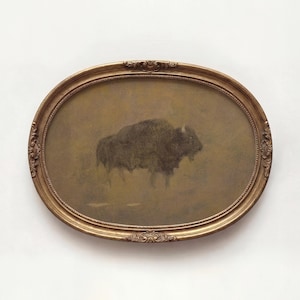 May include: An oval-shaped framed artwork featuring a bison. The artwork is in muted tones of brown and tan, with the bison depicted in the center. The frame is gold-colored with ornate detailing.