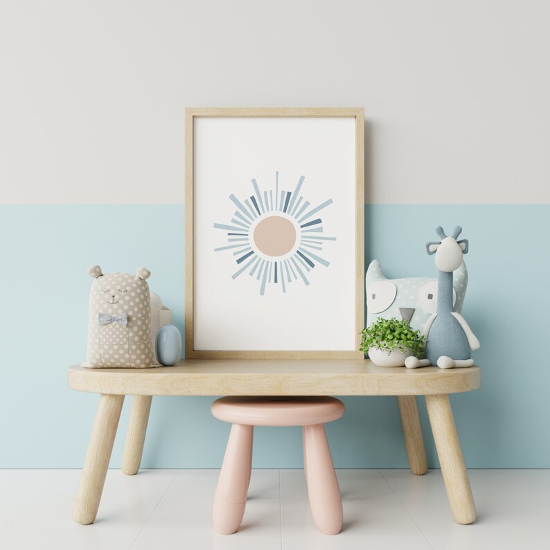 May include: A framed art print featuring a sunburst design in beige, light blue, and navy blue. The artwork is displayed on a light wood table with a pink stool. Soft toys, including a bear and a giraffe, are arranged around the print, creating a nursery or children's room theme.