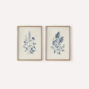 May include: Two framed botanical prints. Each print features a detailed illustration of blue flowers and foliage against a cream-colored background. The frames are a warm gold color, adding a touch of elegance to the artwork. The prints are a set, perfect for adding a touch of nature to any room.
