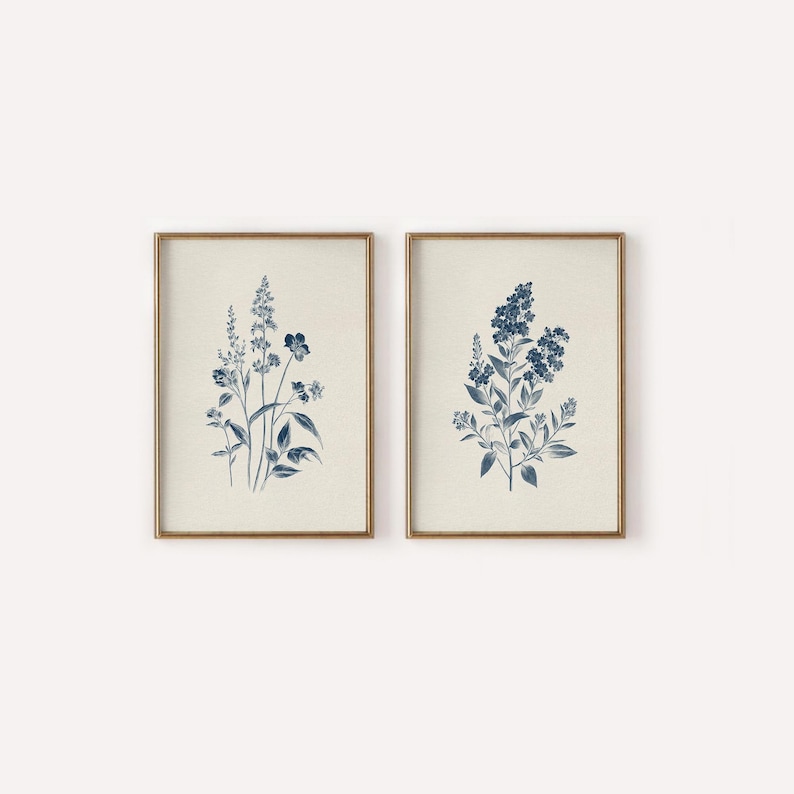 May include: Two framed botanical prints. Each print features a detailed illustration of blue flowers and foliage against a cream-colored background. The frames are a warm gold color, adding a touch of elegance to the artwork. The prints are a set, perfect for adding a touch of nature to any room.