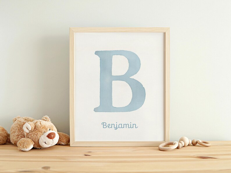 May include: A framed print with a light blue letter "B" and the name "Benjamin" beneath. The frame is light wood. A teddy bear and a wooden rattle sit on a wooden surface in front of the print. The print is in a light wood frame.