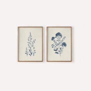 May include: Two framed botanical prints. Each print features a detailed blue floral illustration on a cream background. The frames are a warm gold color, adding a touch of elegance to the artwork. The prints are a matching set.