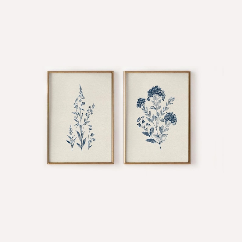 May include: Two framed botanical prints. Each print features a detailed blue floral illustration on a cream background. The frames are a warm gold color, adding a touch of elegance to the artwork. The prints are a matching set.