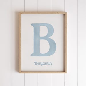 May include: A framed print with a light blue watercolour letter 'B' on a white background. The name 'Benjamin' is written in a matching blue font below the letter. The frame is made of light-coloured wood.