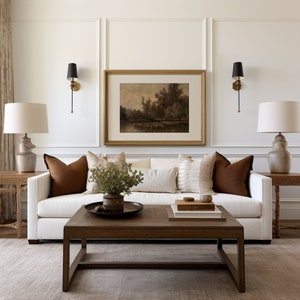 May include: A living room scene featuring a white sofa with brown accent pillows, a wooden coffee table, and a framed landscape painting. Two lamps with white shades flank the sofa. The room has a neutral color palette with a rug and wall sconces.