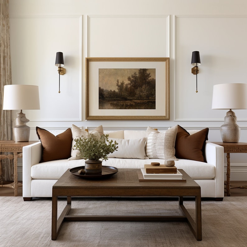 May include: A living room scene featuring a white sofa with brown accent pillows, a wooden coffee table, and a framed landscape painting. Two lamps with white shades flank the sofa. The room has a neutral color palette with a rug and wall sconces.