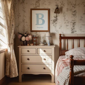 May include: A bedroom interior with a framed print displaying a blue letter "B" and the name "Benjamin". A cream chest of drawers holds a lamp and flowers. The room features floral wallpaper and a wooden bed frame. The scene evokes a cosy atmosphere.