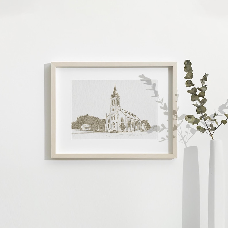 May include: A framed print featuring a detailed sketch of a church with a tall steeple. The artwork is in a neutral color palette, set within a light-colored wooden frame. The image is displayed on a white wall, with a vase of greenery to the right.
