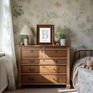 May include: A wooden chest of drawers with six drawers, a framed print with a blue letter "B", and a table lamp. The room has floral wallpaper and a bed with a floral patterned blanket. A teddy bear sits on the chest of drawers.