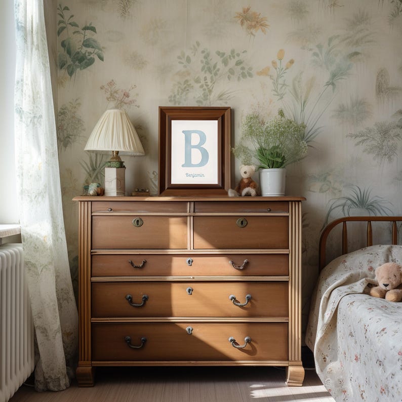 May include: A wooden chest of drawers with six drawers, a framed print with a blue letter "B", and a table lamp. The room has floral wallpaper and a bed with a floral patterned blanket. A teddy bear sits on the chest of drawers.