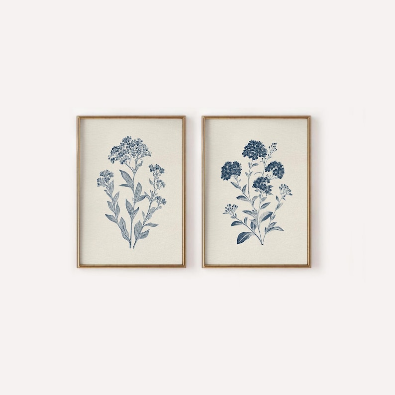 May include: Two framed botanical prints. Each print features a blue floral illustration on a cream background. The flowers have detailed leaves and blooms. The frames are a warm gold color, adding a touch of elegance to the artwork.