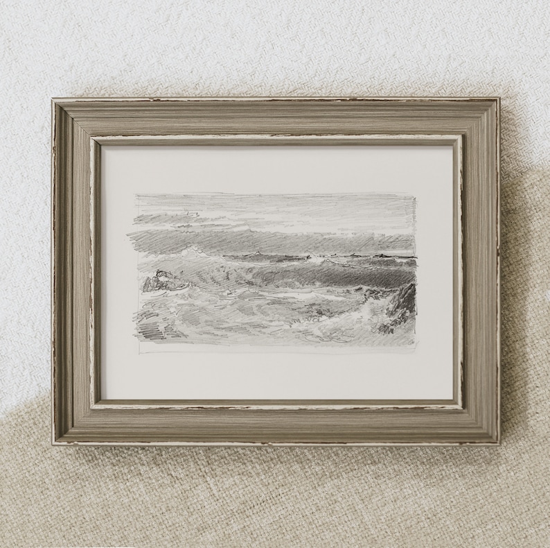 May include: A framed black and white pencil drawing of a seascape. The drawing depicts a rocky coastline with waves crashing against the shore. The frame is a light brown wood with a distressed finish.