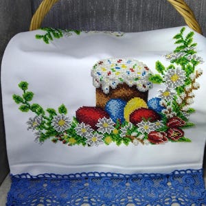 Op de afbeelding: Een witte stoffen mandhoes met een kralen kruissteekontwerp van een cilindrische cake met wit glazuur en kleurrijke hagelslag, omringd door rode en blauwe eieren, witte madeliefjes en groen gebladerte, afgewerkt met een blauwe kanten rand.