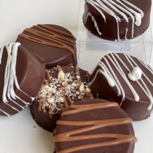 May include: A close-up of six chocolate-covered cookies with different toppings. Some cookies have white chocolate drizzle, others have a sprinkle of nuts, and one has a swirl of milk chocolate.