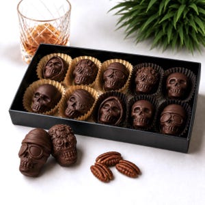 May include: A black rectangular box containing ten dark chocolate skulls in individual paper liners, with two additional chocolate skulls and three pecan nuts placed in front of the box. The skulls feature various designs including goggles and floral patterns.