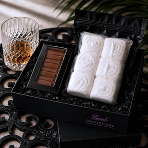 May include: A black gift box containing a clear plastic pouch of six white square marshmallows with spiral patterns and a rectangular black box with a window revealing a brown chocolate bar, next to a crystal glass filled with amber liquid.