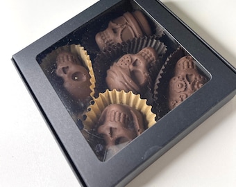Könnte beinhalten: Eine schwarze Box mit einem transparenten Fenster, das sechs schokoladenförmige Schädelbonbons zeigt. Die Bonbons sind in einem Kreis angeordnet, wobei die Schädel nach außen zeigen.