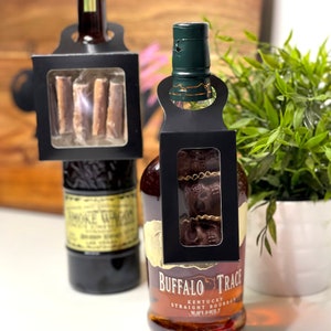 May include: Two bottles of liquor with black gift tags. The tag on the left bottle has a window showing three chocolate bars. The tag on the right bottle has a window showing three chocolate skulls. The bottle on the right is Buffalo Trace Kentucky Straight Bourbon Whiskey.
